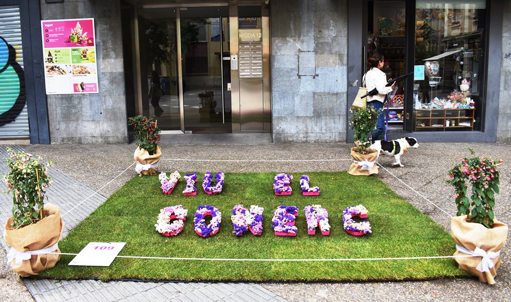 Un quadrat de gespa al terra i sobre un text amb flors que diu: viu el comerç
